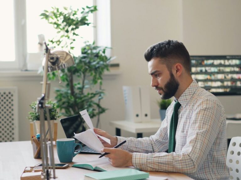 Man at an office desk reviewing printed financial documents with a pen beside a laptop