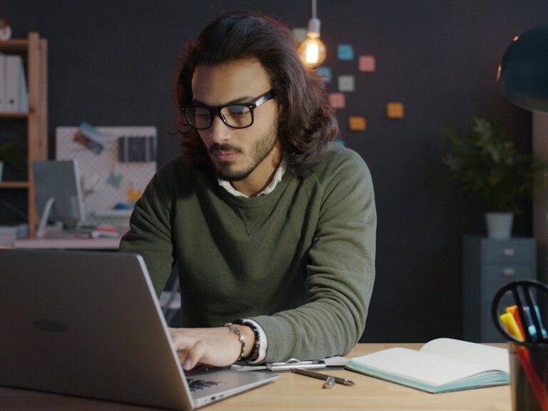 Man in glasses working on a laptop at a desk in a dimly lit office in the evening