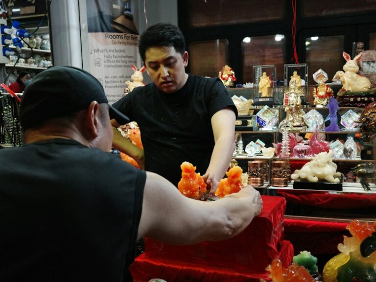Two men arranging orange jade lion figurines at a small shop counter in Singapore's Chinatown