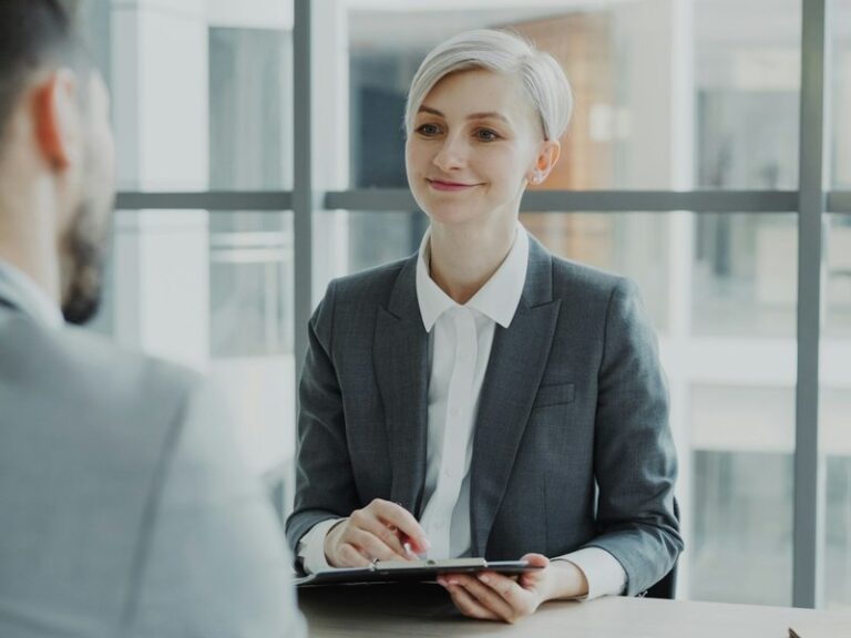 HR interviewer in a grey suit holding a clipboard during a meeting with a male candidate across a desk