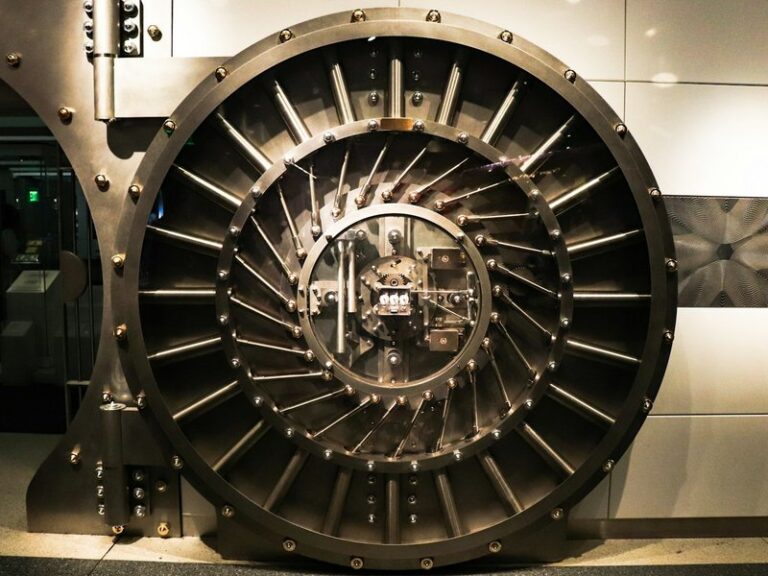 Round steel bank vault door with radial locking bolts, set into a light-coloured wall