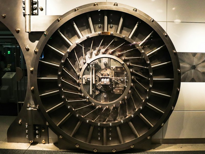 Round steel bank vault door with radial locking bolts, set into a light-coloured wall