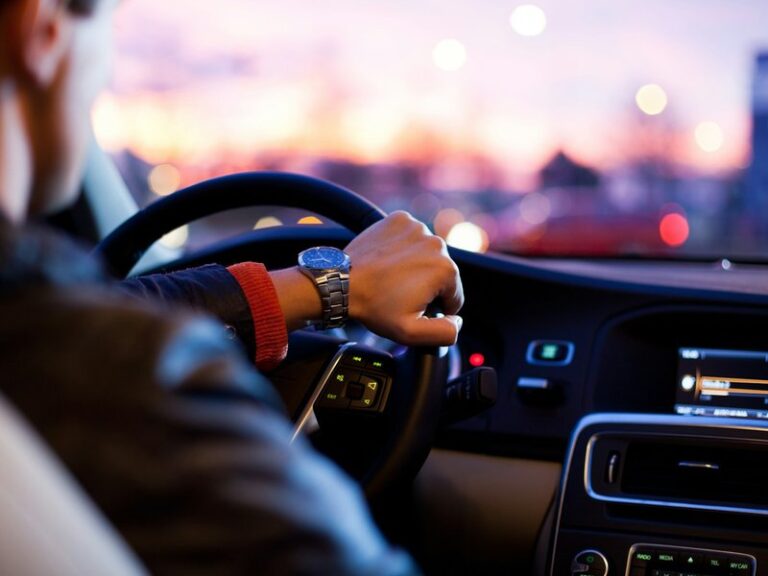 Driver's hand on the steering wheel of a car at dusk, with a city skyline blurred through the windscreen