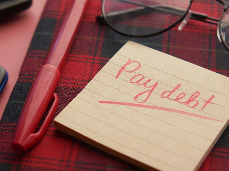 Handwritten sticky note reading Pay debt beside a red pen and reading glasses on a tartan cloth