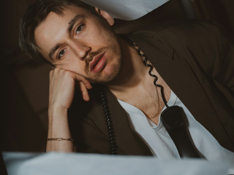 A man in a jacket rests his head on his hand beside a telephone and loose papers, looking dejected