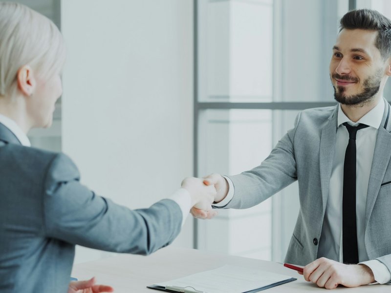 Two professionals in grey suits shaking hands across an office desk with a clipboard