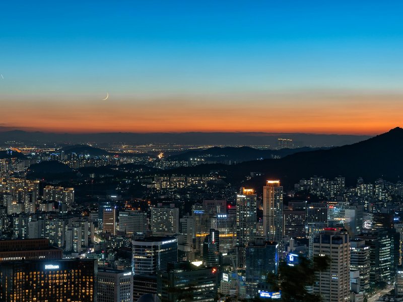 Seoul city skyline at dusk with a crescent moon above the mountains