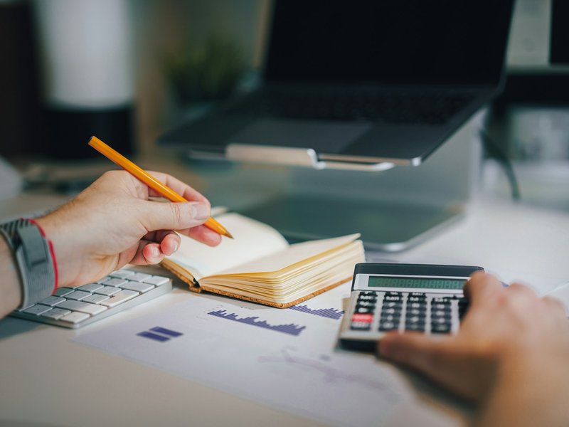 Hands using a calculator and writing in a notebook beside a laptop and printed charts on a desk
