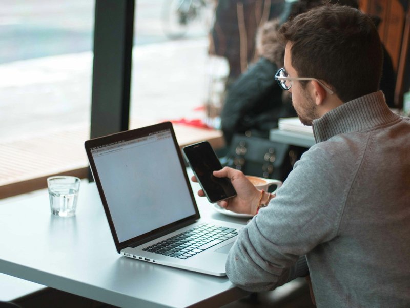Man working on a laptop and smartphone at a cafe table by a window