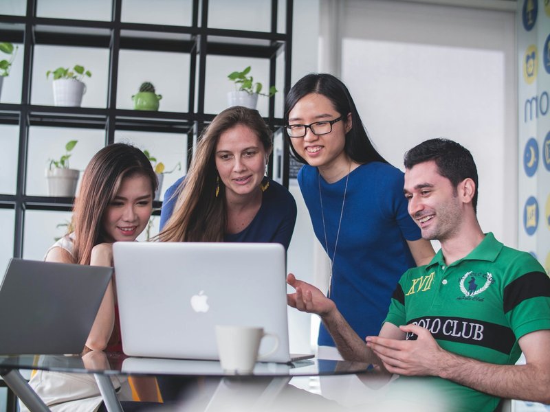 Four young colleagues gathered around a laptop in a bright office, smiling as they review work together