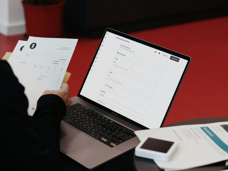 Person holding printed invoices beside a laptop showing an invoice editing screen
