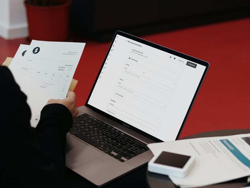 Person holding printed invoices beside a laptop showing an invoice editing screen