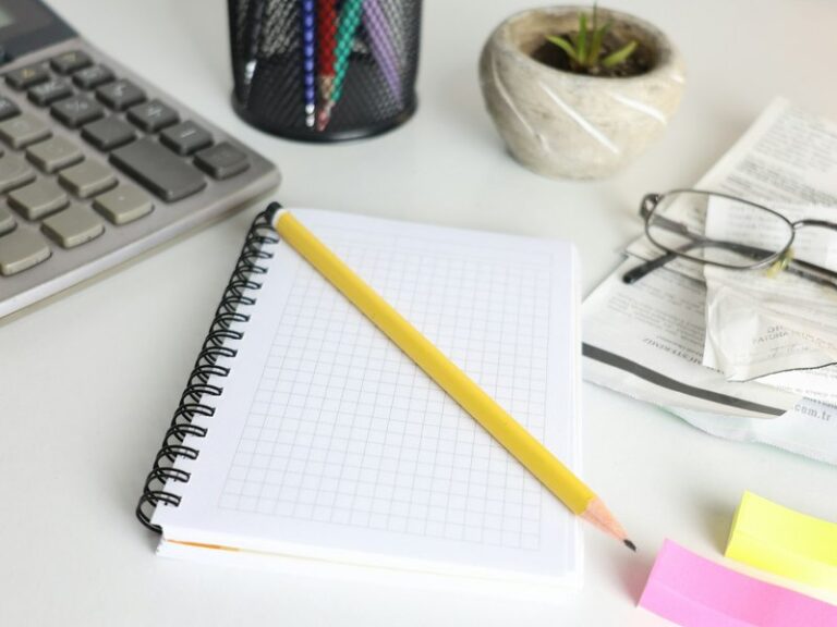 Desk with a calculator, a grid notebook and pencil, reading glasses on folded receipts and sticky notes