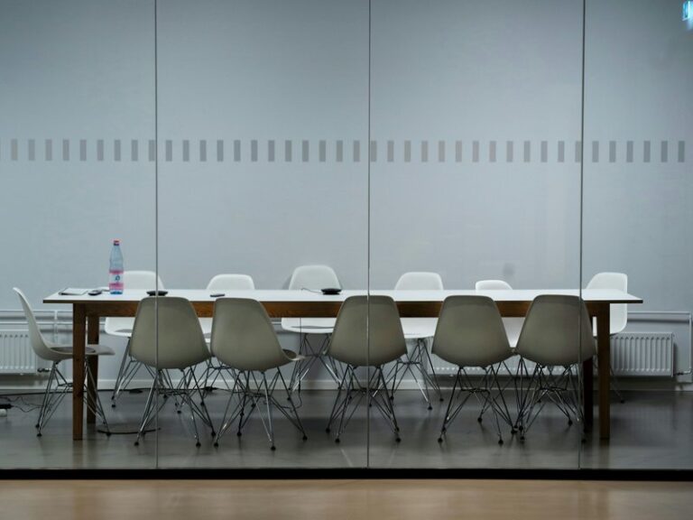 Empty meeting room with a long white table and moulded chairs behind a glass wall, the kind of room where creditors meet to vote on a judicial manager's proposals