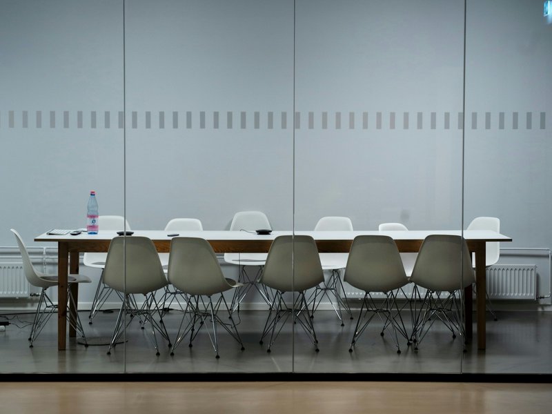 Empty meeting room with a long white table and moulded chairs behind a glass wall, the kind of room where creditors meet to vote on a judicial manager's proposals