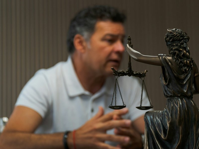 Statue of Lady Justice holding scales in the foreground, with a man in a white polo shirt speaking at a table behind it