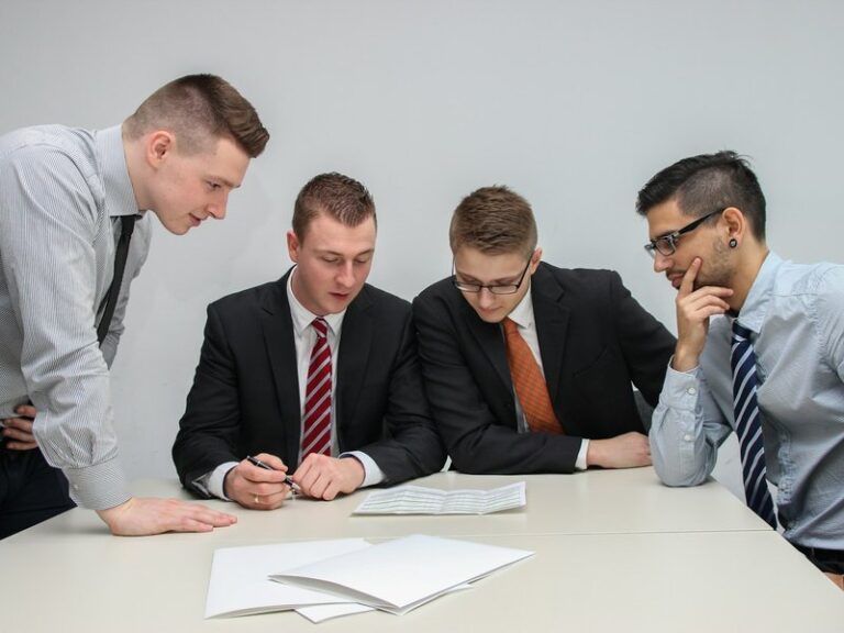Four men in shirts and ties reviewing documents together at a white meeting table