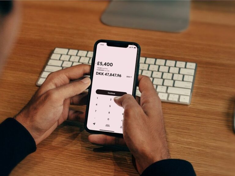 Hands holding a smartphone showing a money transfer screen above a keyboard on a wooden desk