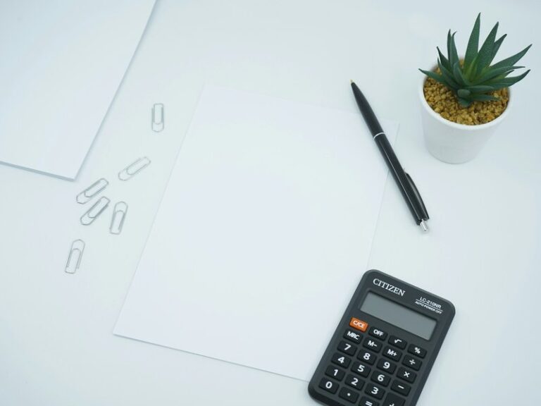 Black calculator and pen beside a blank sheet of paper on a white desk, as used to work out interest on an overdue invoice