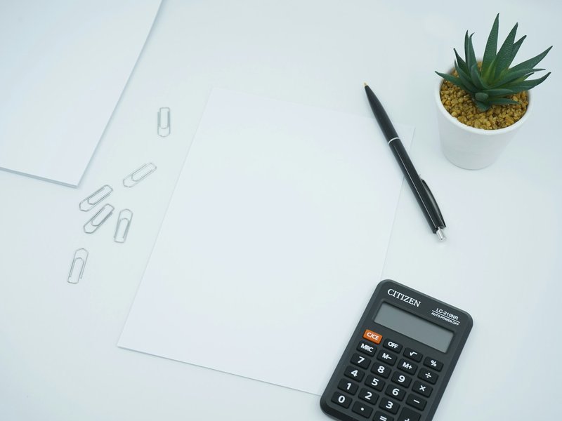 Black calculator and pen beside a blank sheet of paper on a white desk, as used to work out interest on an overdue invoice