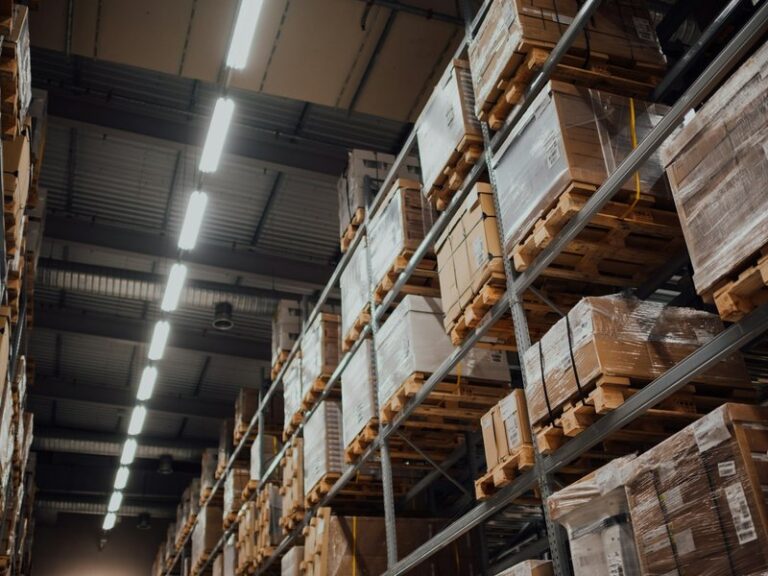 Warehouse racks stacked with pallets of boxed stock under strip lighting