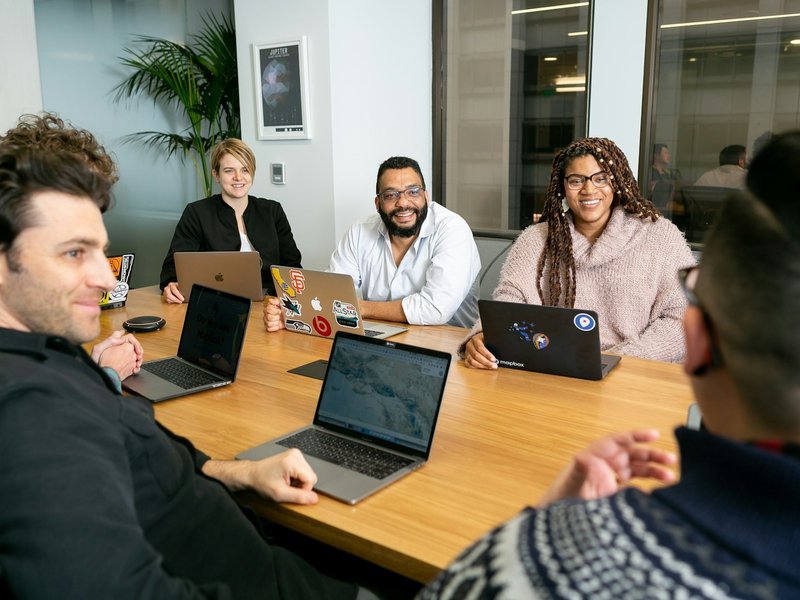 Five colleagues with laptops around a wooden table in an office meeting room