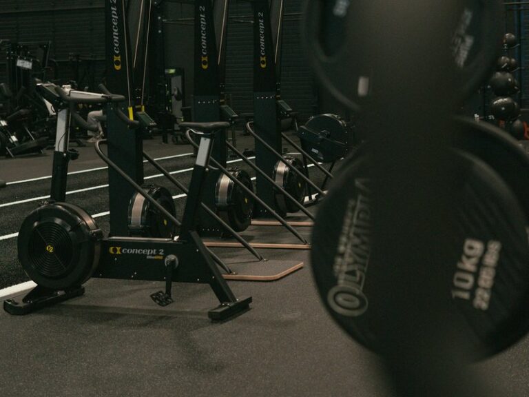 Air bikes and ski ergometers on a gym floor with 10 kg weight plates in the foreground