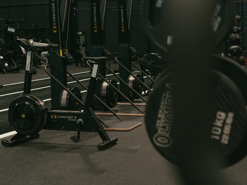 Air bikes and ski ergometers on a gym floor with 10 kg weight plates in the foreground