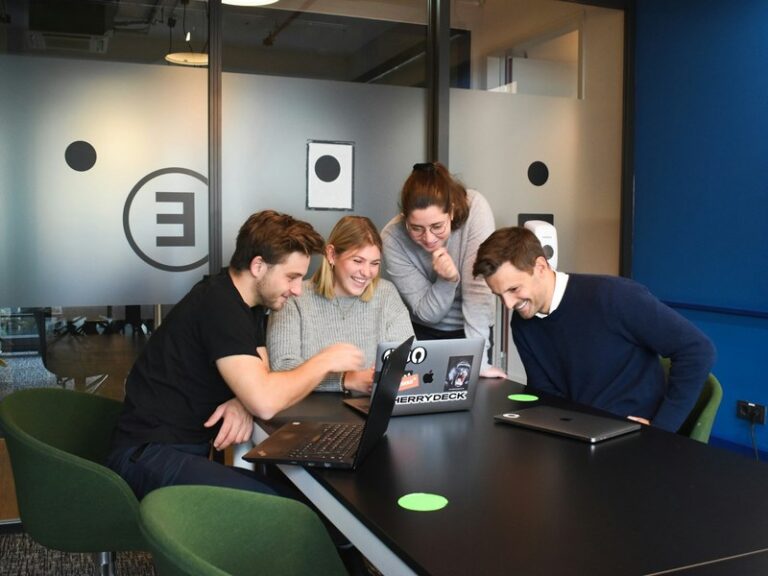 Four agency colleagues gathered around a laptop at a meeting table, smiling at a campaign on screen