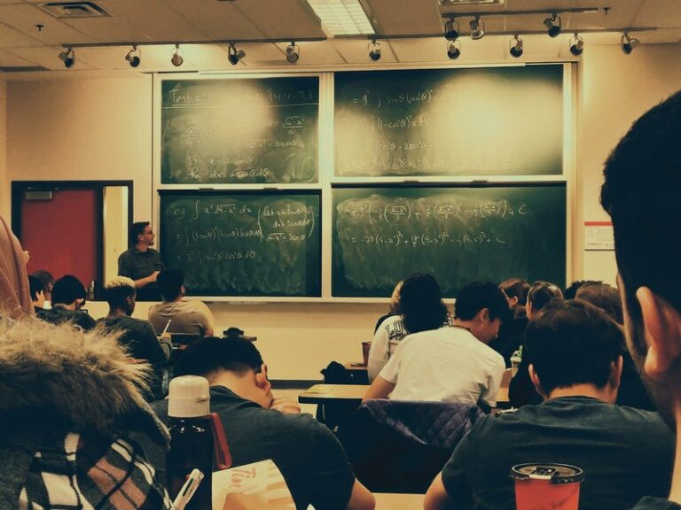 Students seated in a classroom facing a teacher who stands beside chalkboards covered in maths equations