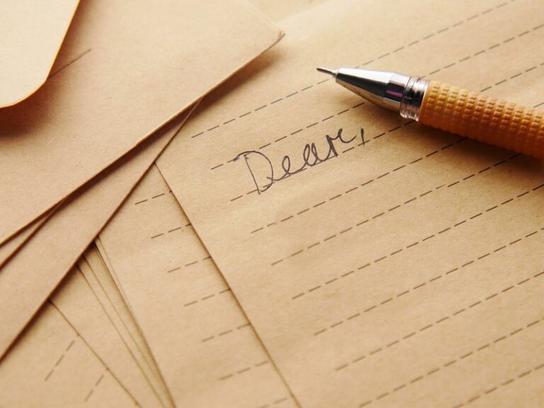 Handwritten letter beginning "Dear" on brown lined paper beside envelopes and a pen