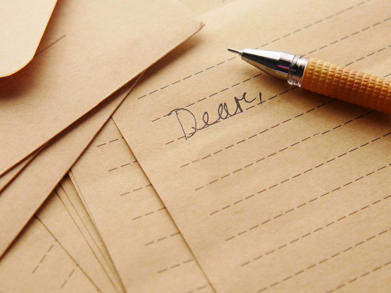 Handwritten letter beginning "Dear" on brown lined paper beside envelopes and a pen