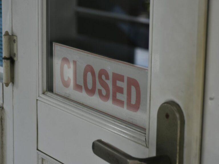 A red CLOSED sign behind the glass panel of a white shop door with a metal handle