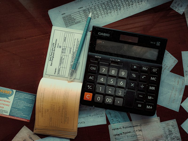 Calculator, pen and receipt book on a wooden desk surrounded by loose receipts
